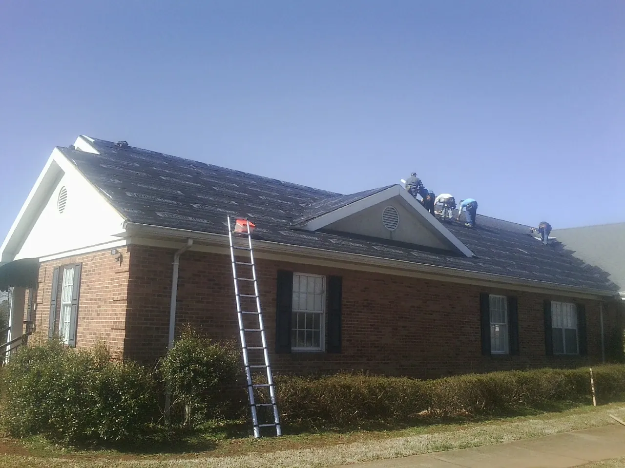 Crew tearing shingles off a brick ranch roof with a ladder set — Emergency Roof Repair work by our crew in Gill