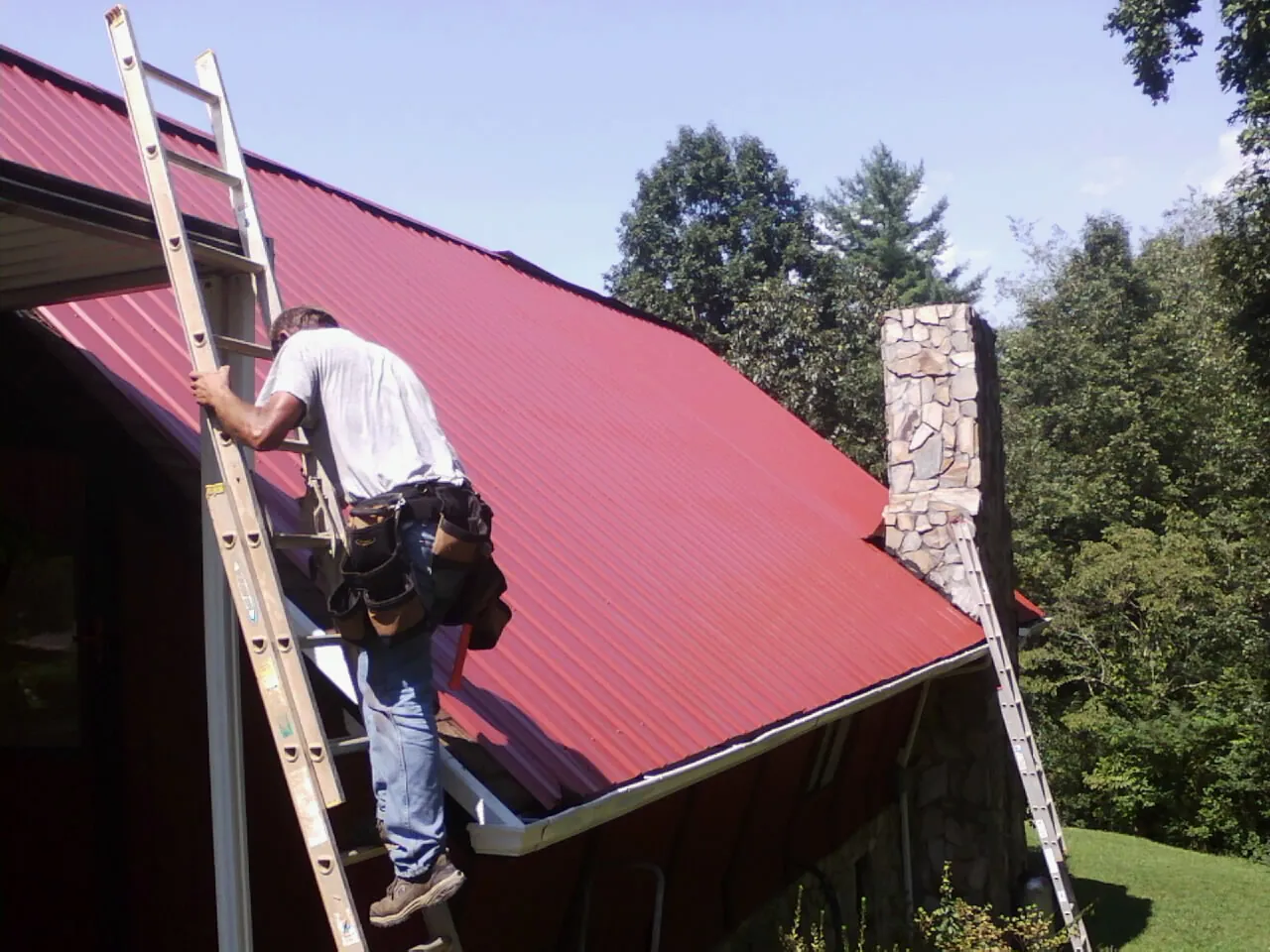Roofer on a ladder beside a red metal roof and stone chimney — Soffit & Fascia Repair by Roofing Brothers in Gill